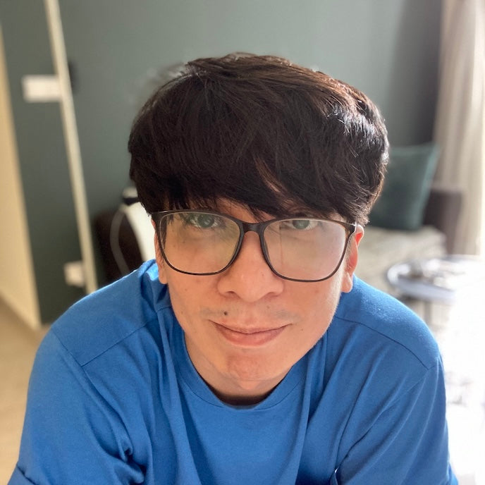 A close-up portrait of a man with dark hair and glasses, wearing a blue t-shirt in an indoor setting.
